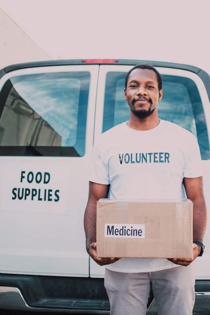 Volunteer holding box of medicine in front of food supply van, showcasing community aid.