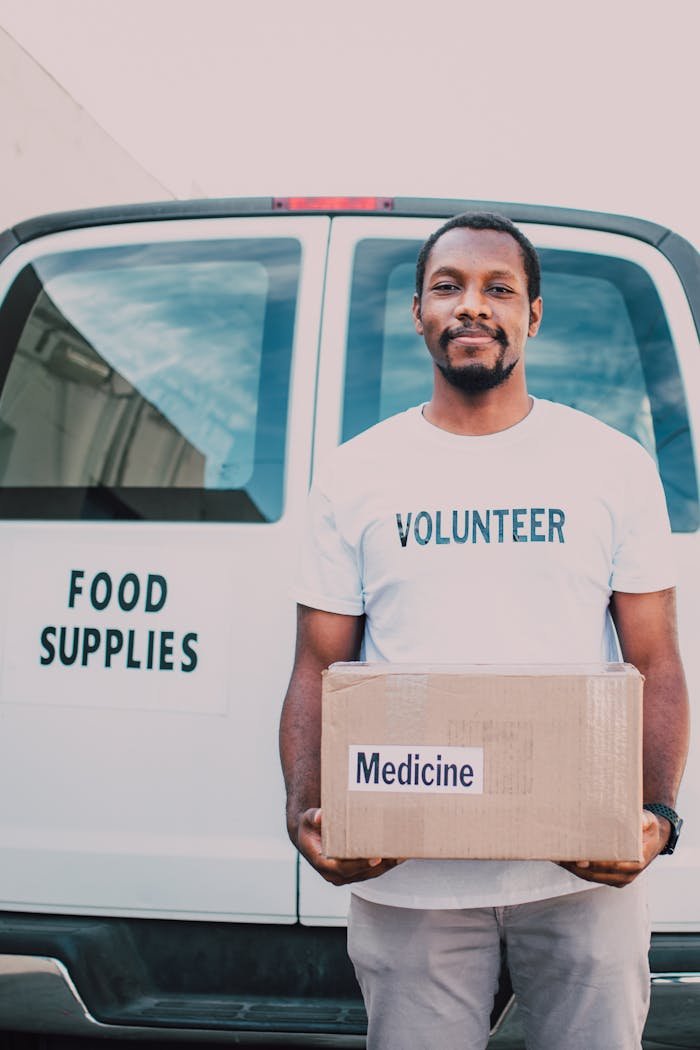 Volunteer holding box of medicine in front of food supply van, showcasing community aid.