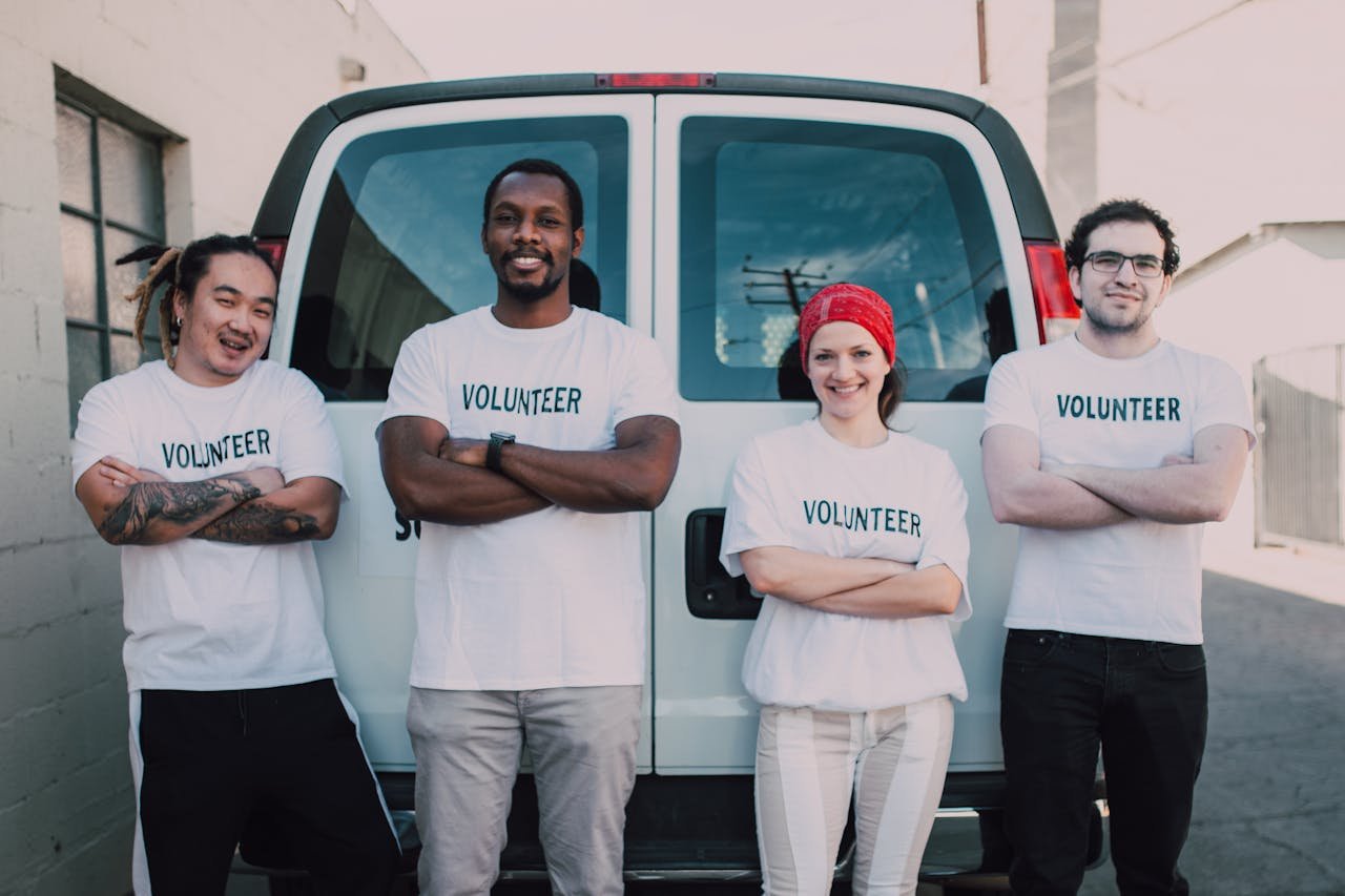 Diverse group of adult volunteers smiling in front of a van during a sunny day.